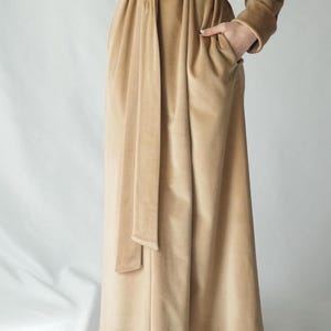 Pu&ograve; includere: Lungo abito in velluto beige con cintura abbinata. L'abito ha maniche lunghe e tasche. Il tessuto ha un aspetto morbido e lussuoso. L'abito &egrave; di colore neutro, adatto a varie occasioni.