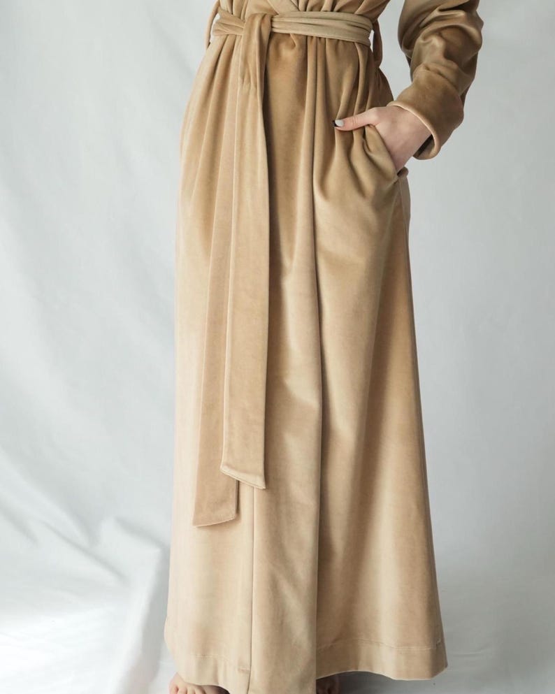 Pu&ograve; includere: Lungo abito in velluto beige con cintura abbinata. L'abito ha maniche lunghe e tasche. Il tessuto ha un aspetto morbido e lussuoso. L'abito &egrave; di colore neutro, adatto a varie occasioni.