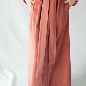 May include: A full-length, coral-colored velvet robe with long sleeves and a matching belt. The robe has pockets and is tied at the waist. The fabric has a soft, luxurious appearance.