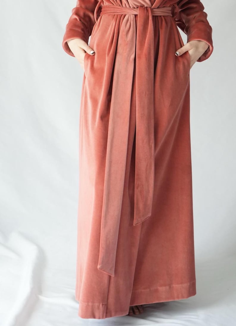 May include: A full-length, coral-colored velvet robe with long sleeves and a matching belt. The robe has pockets and is tied at the waist. The fabric has a soft, luxurious appearance.