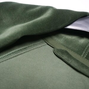 Pu&ograve; includere: Primo piano di un capo in velluto verde, forse una giacca o un cappotto. Il tessuto ha una consistenza morbida e soffice. Il colletto e il risvolto sono visibili, con una consistenza a contrasto. Il capo &egrave; appeso a una gruccia nera.