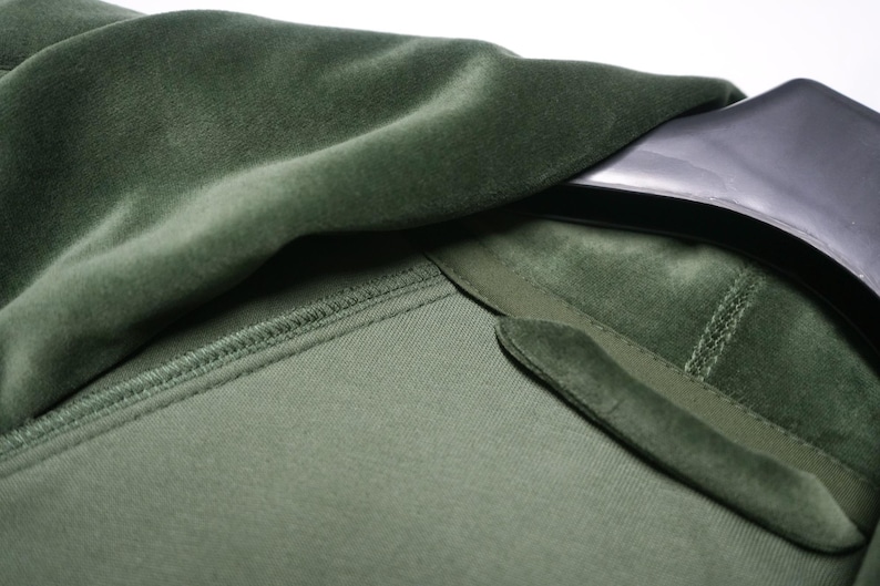 Pu&ograve; includere: Primo piano di un capo in velluto verde, forse una giacca o un cappotto. Il tessuto ha una consistenza morbida e soffice. Il colletto e il risvolto sono visibili, con una consistenza a contrasto. Il capo &egrave; appeso a una gruccia nera.
