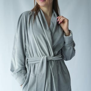 Pu&ograve; includere: Un accappatoio grigio chiaro, in tessuto felpato, con collo a scialle e cintura abbinata annodata in vita. L'accappatoio ha maniche lunghe e tasche. Il tessuto appare morbido e confortevole, adatto al relax. Lo stile generale &egrave; semplice ed elegante.