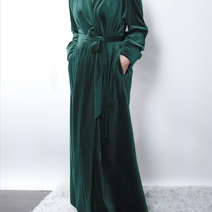 May include: A long, emerald green velvet robe with a shawl collar, long sleeves, and a matching waist tie. The robe has two front pockets and is floor-length. The fabric has a soft, luxurious appearance.