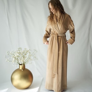 May include: A long, tan robe with a matching belt is shown. The robe has long sleeves and a shawl collar. A gold spherical vase with white flowers is on the left. The robe appears to be made of a soft, velvety material.