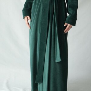 May include: A long, emerald green velvet robe with a matching belt tied at the waist. The robe has long sleeves with folded cuffs. The fabric has a soft, luxurious appearance.