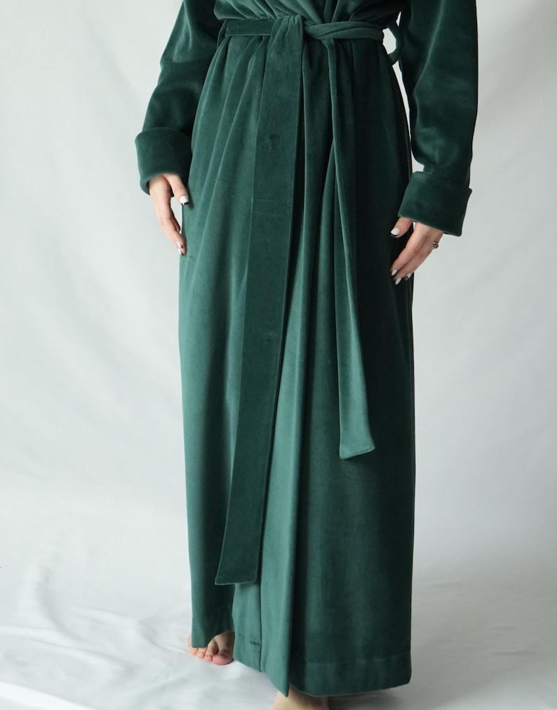 May include: A long, emerald green velvet robe with a matching belt tied at the waist. The robe has long sleeves with folded cuffs. The fabric has a soft, luxurious appearance.