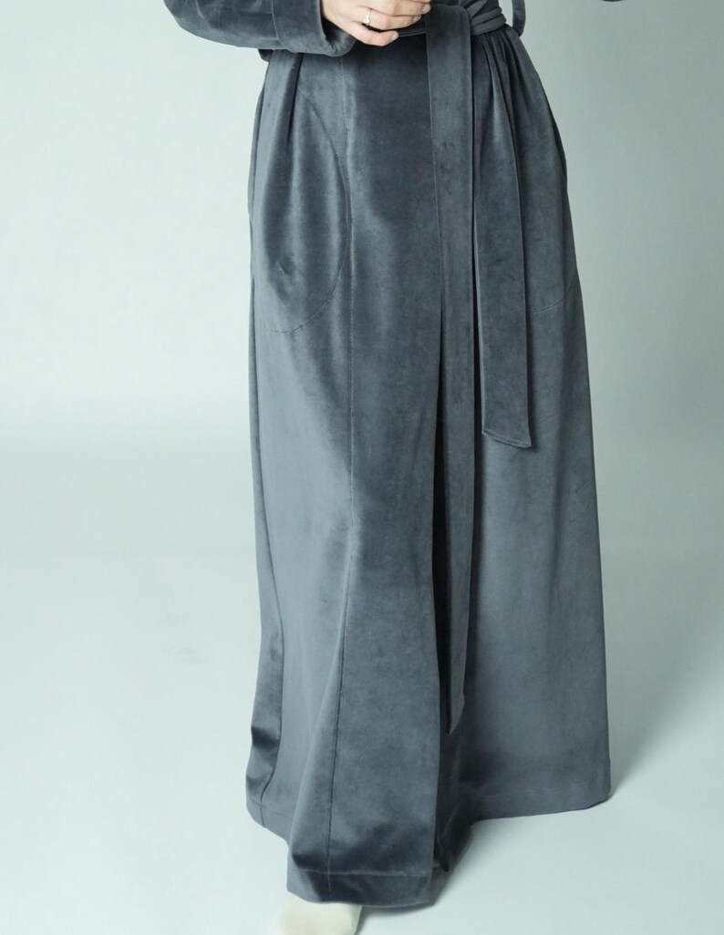May include: A long, flowing, gray velvet robe with a matching belt. The robe has a soft, luxurious appearance and features a wrap-around design. The fabric drapes elegantly, suggesting comfort and style. The robe is suitable for lounging or as a stylish layering piece.