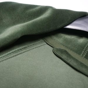May include: Close-up of a green velvet garment, possibly a jacket or coat. The fabric has a soft, plush texture. The collar and lapel are visible, with a contrasting texture. The garment is hanging on a black hanger.