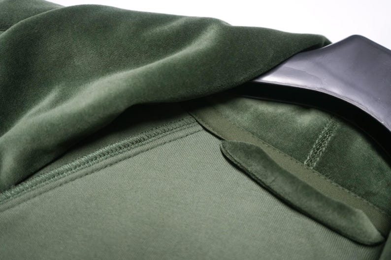May include: Close-up of a green velvet garment, possibly a jacket or coat. The fabric has a soft, plush texture. The collar and lapel are visible, with a contrasting texture. The garment is hanging on a black hanger.