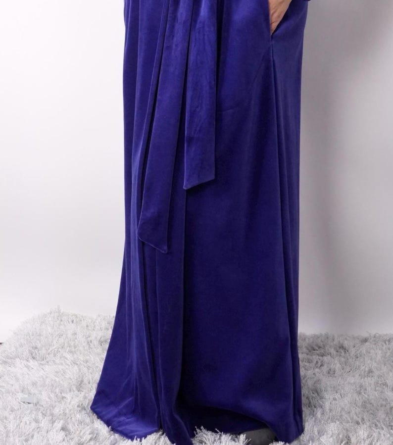 Pu&ograve; includere: Un lungo abito blu reale con una cintura in tessuto abbinata annodata in vita. L'abito presenta un design drappeggiato e una lucentezza sottile, che suggerisce un tessuto morbido e lussuoso. L'abito, lungo fino al pavimento, sembra essere realizzato in velluto o materiale simile.