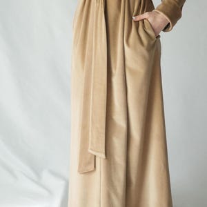 May include: A long, tan velvet robe with a matching belt and pockets. The robe has long sleeves and a flowing skirt. The model is wearing the robe with their hand in the pocket. The robe is made of a soft, luxurious fabric.