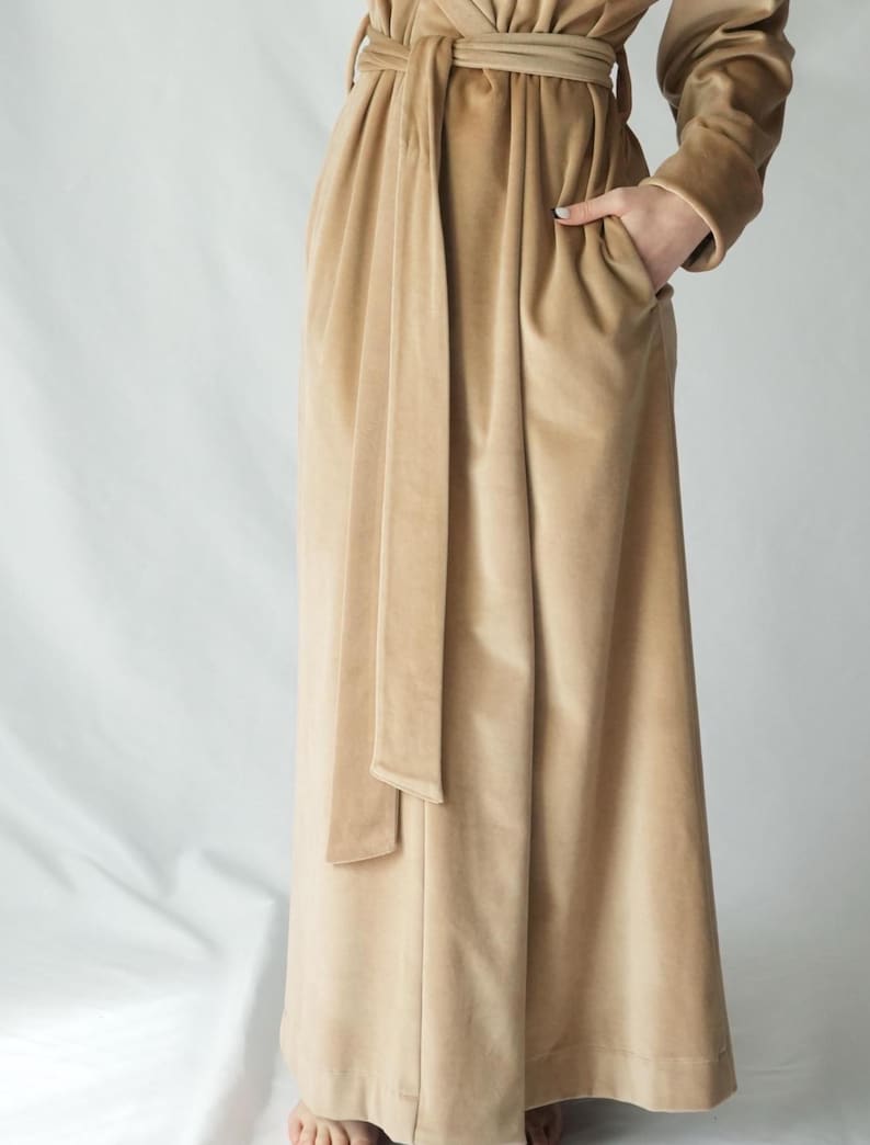 May include: A long, tan velvet robe with a matching belt and pockets. The robe has long sleeves and a flowing skirt. The model is wearing the robe with their hand in the pocket. The robe is made of a soft, luxurious fabric.