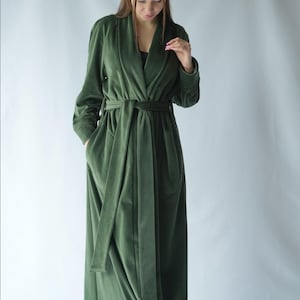 Pu&ograve; includere: Un lungo accappatoio in velluto verde oliva con collo a scialle e cintura abbinata. L'accappatoio ha maniche lunghe e tasche. La modella indossa un top nero sotto. L'accappatoio &egrave; lungo fino al pavimento.
