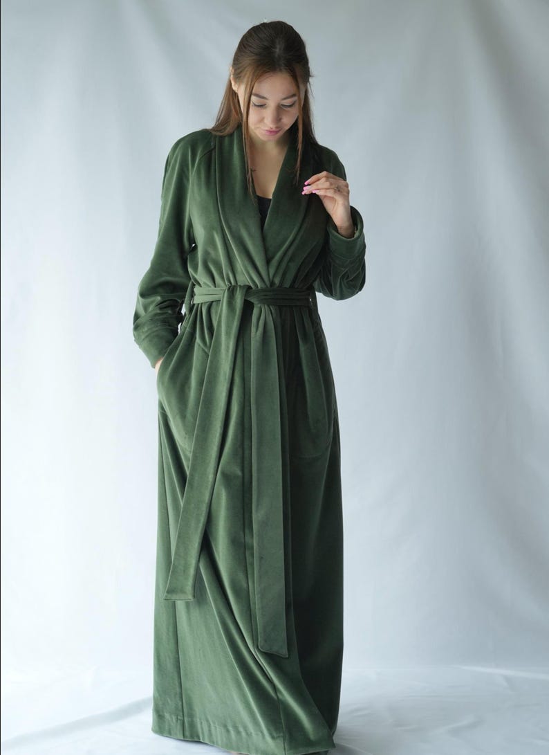 Pu&ograve; includere: Un lungo accappatoio in velluto verde oliva con collo a scialle e cintura abbinata. L'accappatoio ha maniche lunghe e tasche. La modella indossa un top nero sotto. L'accappatoio &egrave; lungo fino al pavimento.