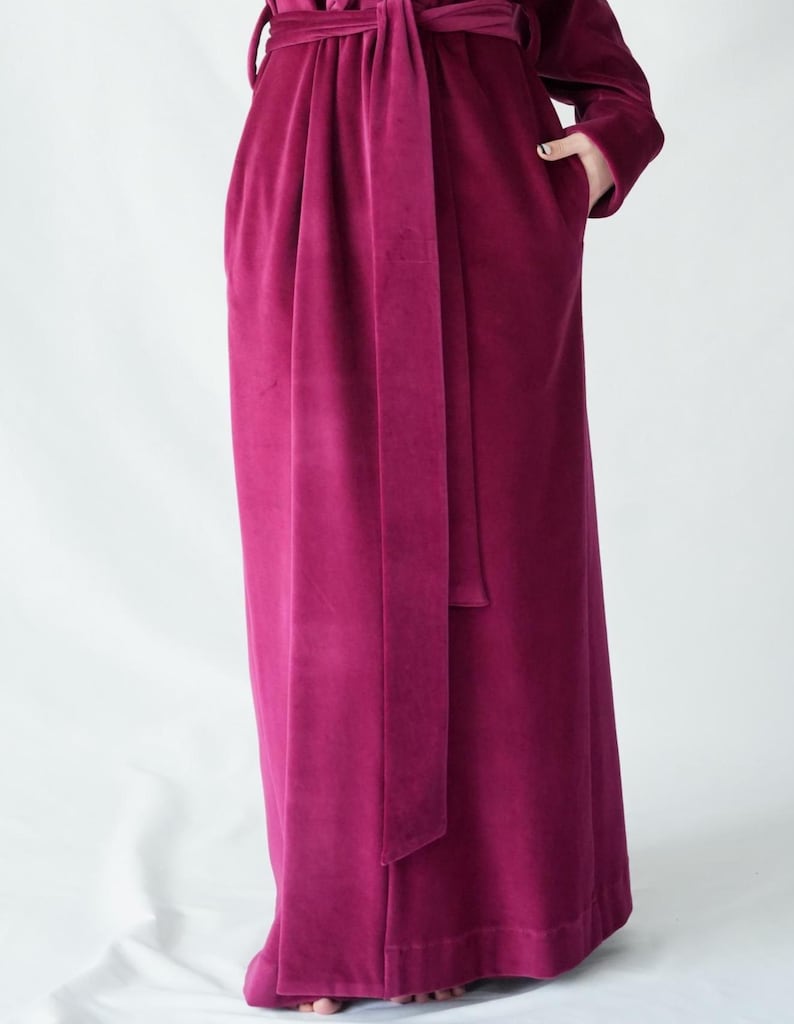 May include: A long, magenta velvet robe with a matching belt tied at the waist. The robe has long sleeves and pockets. The fabric has a soft, plush texture.