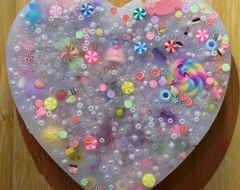 May include: A heart-shaped resin art piece filled with colorful candies, beads, and small decorative elements. The design includes a variety of sweets, such as lollipops, candies, and sprinkles, in a mix of pastel colors. The piece is set on a wooden surface.