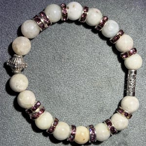 May include: A beaded bracelet featuring round, off-white beads interspersed with silver-toned spacers adorned with small, purple gemstones. The bracelet includes decorative silver-toned end pieces.