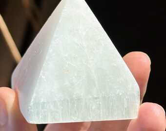Puede incluir: Una pirámide de selenita verde claro, sostenida en una mano. La pirámide tiene una superficie lisa y pulida y una forma triangular. El cristal es translúcido. La pirámide mide aproximadamente 7.6 cm de altura.