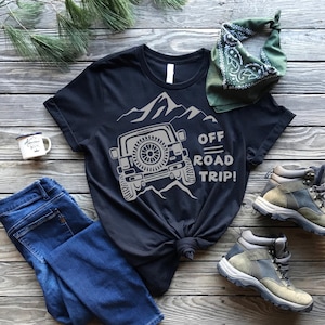 Puede incluir: Camiseta negra con un gráfico blanco de un jeep conduciendo por las montañas. El texto "Off Road Trip!" está impreso en la camiseta.