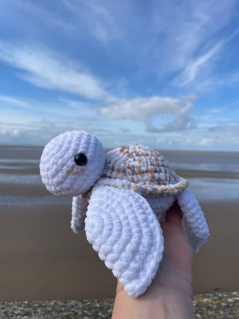Op de afbeelding: Een witte gehaakte schildpad met een bruin en witte schild. De schildpad wordt in een hand gehouden tegen een blauwe lucht achtergrond.