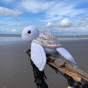 Op de afbeelding: Wit gebreid schildpad speelgoed met een bruin en witte schelp die op een stuk drijfhout met zeewier zit.