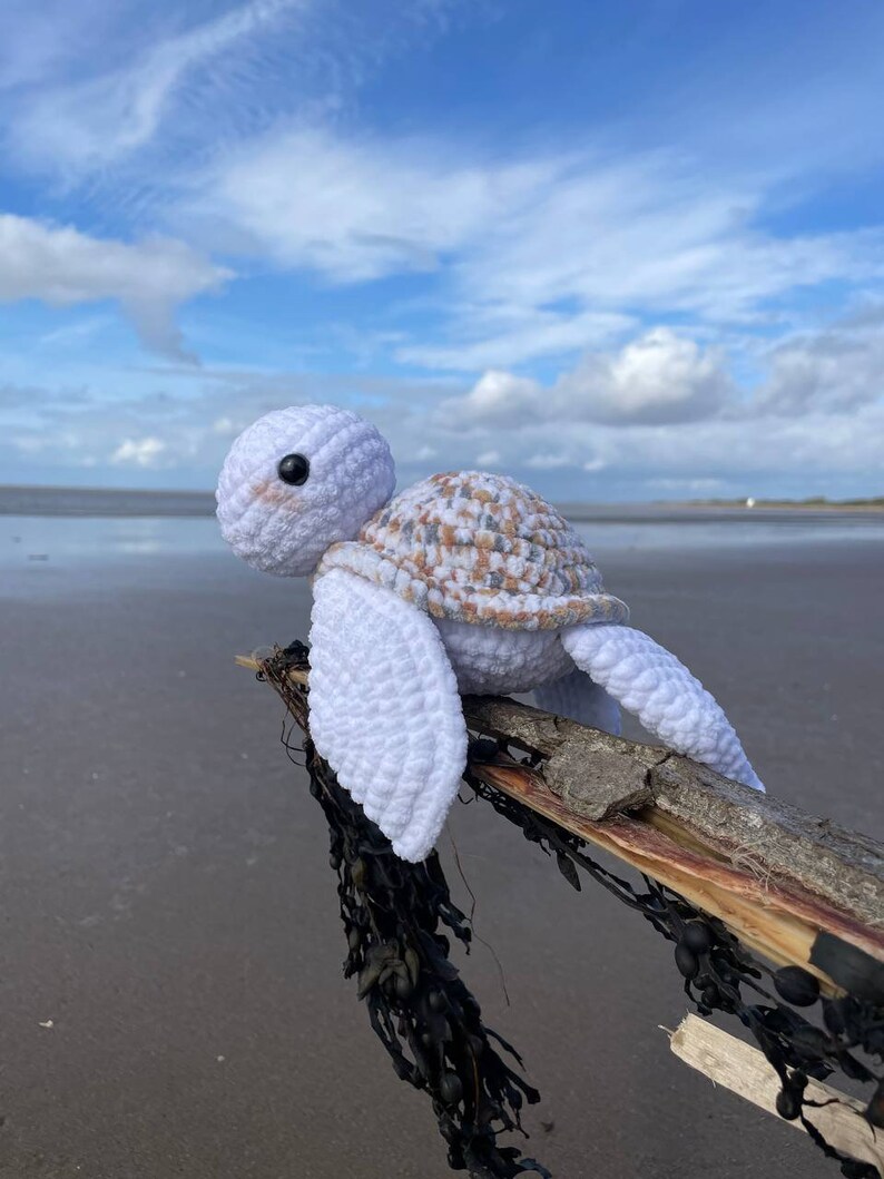 Op de afbeelding: Wit gebreid schildpad speelgoed met een bruin en witte schelp die op een stuk drijfhout met zeewier zit.