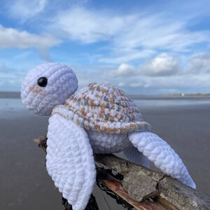 Op de afbeelding: Een witte gehaakte schildpad met een bruine en beige schild zit op een tak van drijfhout. De schildpad heeft zwarte ogen en een witte achtergrond.