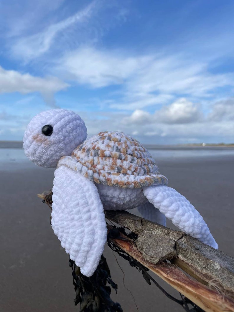 Op de afbeelding: Een witte gehaakte schildpad met een bruine en beige schild zit op een tak van drijfhout. De schildpad heeft zwarte ogen en een witte achtergrond.