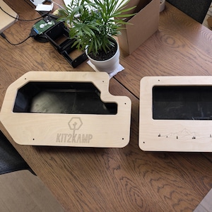 May include: Two wooden storage containers with black interiors. One has a unique shape with the text "KIT2KAMP" and a logo. The other has a rectangular shape with a mountain and tree design. Both are on a wooden table.