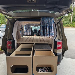 Puede incluir: Una conversión interior de furgoneta con una estructura de cama de madera, cajones de almacenamiento y cortinas. Los cajones abiertos revelan espacio de almacenamiento. Las cortinas tienen un patrón azul y blanco. La puerta trasera de la furgoneta está abierta.