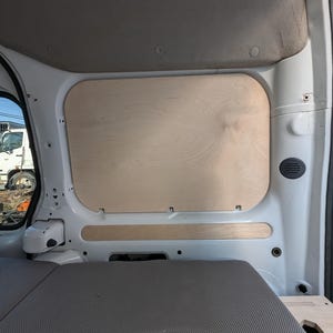 May include: Interior view of a white van with a wooden panel installed on the side wall. The panel is rectangular with rounded corners and a natural wood grain finish. A gray upholstered bench seat is visible.