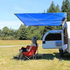 May include: A blue awning extends from a small camper trailer. A person sits in a red folding chair in a grassy field. The trailer is white and gray with a black storage box.