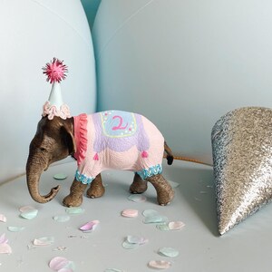 May include: A brown elephant figurine wearing a pink and purple blanket with the number 2 and a small party hat with a pink pom-pom, standing on a round grey box decorated with orange pom-pom trim and a silver glitter party hat nearby.