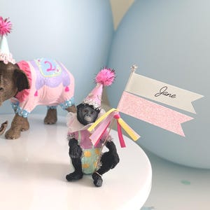 May include: Three animal figurines including a lion, an elephant, and a monkey wearing party hats and colorful outfits. The elephant has a pink blanket with the number 2, and the monkey holds a flag with the name Jane written in black script.