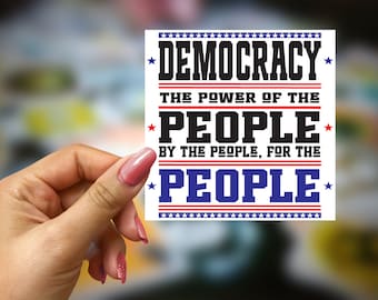 May include: A white sticker with red, white and blue stars and stripes. The sticker says "Democracy - The Power of the People - By the People, For the People".