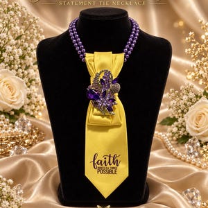 May include: A statement tie necklace featuring a double strand of purple beads and a yellow satin tie. The tie is adorned with a large purple and clear crystal brooch and displays the black text faith MAKES ALL THINGS POSSIBLE.