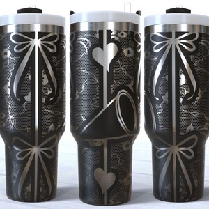 May include: Three stainless steel tumblers with black and silver designs. The tumblers have a black and silver lace pattern with a bow at the top and bottom. The middle tumbler has a heart and a black and silver design.