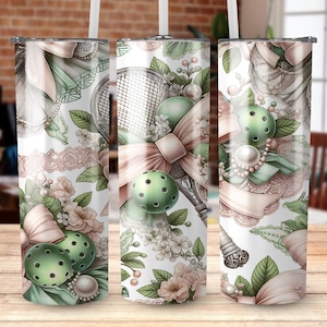 May include: A set of three stainless steel tumblers with a decorative design. The design features a pickleball theme with green pickleballs, a racket, bows, and floral accents in soft pastel colors. The tumblers have a white background.