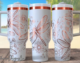 May include: White travel mug with a copper-colored dragonfly and floral design. The mug has a handle and a copper-colored band at the top. The design includes dragonflies, daisies, and stars.