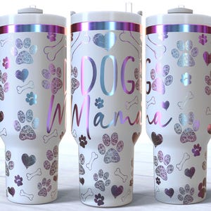 May include: White stainless steel travel mug with a pink and purple iridescent finish. The mug has a handle and a lid. The mug is decorated with a paw print pattern and the text "Dog Mama".