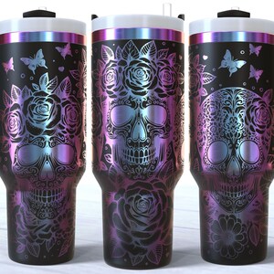 May include: Three stainless steel tumblers with a black background and a design of a sugar skull with flowers and butterflies. The tumblers have a rainbow iridescent finish.