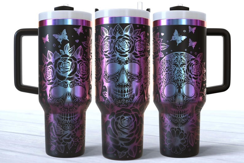 May include: Three stainless steel tumblers with a black background and a design of a sugar skull with flowers and butterflies. The tumblers have a rainbow iridescent finish.