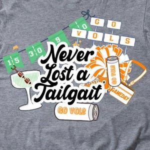 Può includere: T-shirt grigio con un design grafico con la scritta "Never Lost a Tailgate" e illustrazioni a tema tailgate. Il design include un cocktail, una lattina e un biglietto per il giorno della partita.