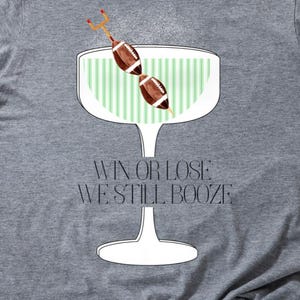 May include: A heather gray t-shirt featuring a cocktail glass design with a football and the text "WIN OR LOSE WE STILL BOOZE". The graphic is in white, green, and brown.