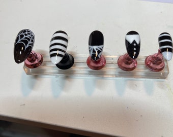 May include: Five black and white nail tips on display stands. The nail tips feature different designs, including a spiderweb, stripes, and a tuxedo.