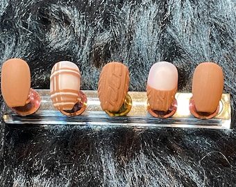 May include: Five brown oval-shaped nail tips with different designs. The designs include a plaid pattern, a textured pattern, a solid color, and a textured pattern with a white accent.