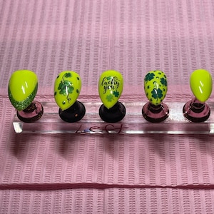 May include: Five green and yellow nail tips with shamrock designs and the text "lucky you" on one of the tips.