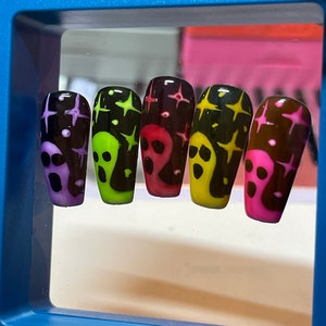 May include: Five fake nails with a black background and white stars. Each nail is a different colour: purple, green, red, yellow, and pink. Each nail has a white ghost face design.