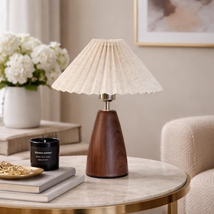 May include: A stylish table lamp featuring a pleated beige lampshade and a dark wood base. The lamp is placed on a marble-topped table with a gold rim, alongside a black scented candle and a vase of white flowers.